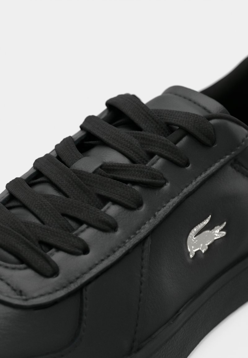 Svart lädersneaker med platta svarta snören, slät yta och silverfärgad logotypaccent på sidan. Enkelt design, låg skärning.