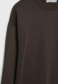 Sweat-shirt brun à manches longues avec col rond en tissu doux, avec une étiquette visible de la marque Stradivarius à l'intérieur du col.
