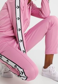 Rosa Trainingsanzug mit schwarzen und weißen Streifenakzenten, versehen mit Sternen und Pfeilen, aus elastischem Material. Weiße Turnschuhe runden das Outfit ab.