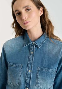 Denim shirt in light blue with two front pockets, a button-up front, and a classic collar. Material shows a faded texture with visible stitching.
