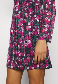 Kukkamekko, jossa on musta tausta monivärisillä kukilla, laskoshuntu, pitkät hihat ja istuva vyötärö. Pehmeä, teksturoitu kangas.