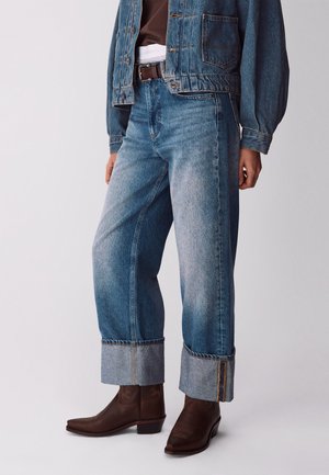 Persona con jeans di jeans blu con risvolto, cintura in pelle marrone, giacca di jeans e stivaletti marrone scuro, in piedi davanti a uno sfondo semplice.