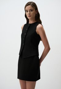 Robe noire ajustée sans manches, avec une fermeture à boutons à l'avant, une texture lisse et une silhouette cintrée, se terminant au-dessus du genou.