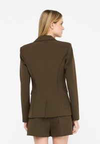 Blazer marrone su misura con spalle strutturate, silhouette aderente e schiena ventilata, abbinato a pantaloni shorts coordinati.