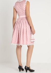 Woman wearing a pink, knee-length, sleeveless dress with white floral patterns, white lace trim, and black Mary Jane heels standing against a white background.