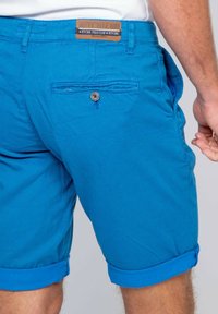 Shorts bleus en tissu texturé, dotés d'une poche arrière avec bouton et d'une étiquette en cuir avec le nom de la marque sur la taille. Ourlets retroussés.