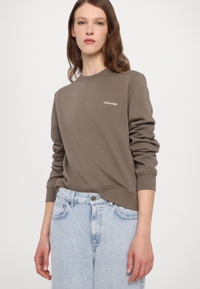 Brun genser med ribbestrikkede mansjetter og nederkant, pyntet med en liten brodert logo på brystet. Kombinert med lyseblå jeans.