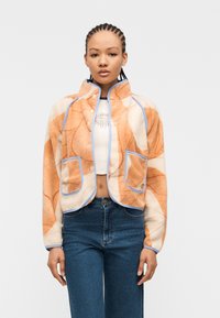 Νεαρή γυναίκα με πλεξούδες cornrow, που φοράει πορτοκαλί και κρεμ fleece μπουφάν με μοτίβο και μπλε περιθώριο, λευκό κοντό τοπ και μπλε τζιν, όρθια.