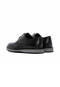 Zwarte leren schoenen met een gestructureerd, geperforeerd oppervlak, buisvormige veters en een platte grijze rubberen zool, met een glad achterpaneel.