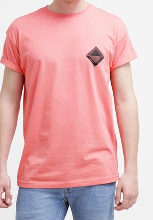 Koralowy t-shirt z krótkim rękawem i podwiniętymi mankietami, z małym czarnym wzorem diamentu na lewej piersi. Miękka, gładka tekstura tkaniny.