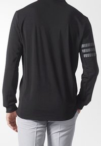 Persona vestida con una chaqueta negra de manga larga con tres franjas grises en la manga izquierda, combinada con pantalones gris claro, de pie con la espalda mostrada.