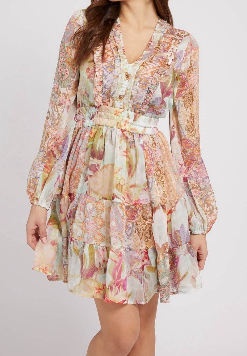 Femme portant une robe fleurie multicolore à manches longues avec des volants et une taille froncée, posant devant un fond uni.