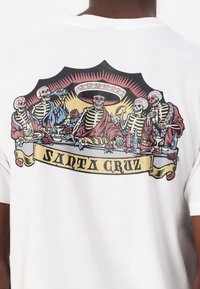 Hvit kortermet t-skjorte med et fargerikt motiv av skjeletter ved et festmåltid, med røde og blå detaljer og et "SANTA CRUZ" banner nederst.