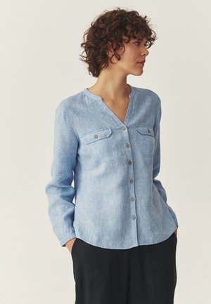 Kobieta o kręconych włosach, w jasnoniebieskiej koszuli zapinanej na guziki z kieszeniami, stojąca z rękami w czarnych kieszeniach spodni, patrząca w prawo