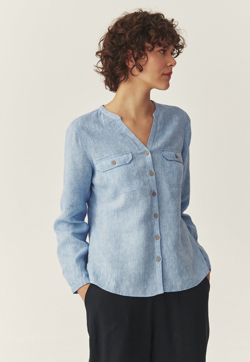 Femme aux cheveux bouclés portant une chemise bleu clair à boutons avec des poches, debout les mains dans les poches d'un pantalon noir, regardant vers la droite