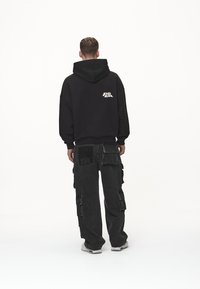 Zwarte sweatvest met capuchon en logo op de achterkant, gecombineerd met losvallende zwarte cargobroek met meerdere zakken en gestructureerde details.