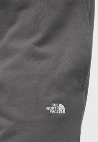 Ύφασμα φούτερ σκούρου γκρι με λεία υφή, που φέρει λευκό κεντητό λογότυπο "The North Face" στο κάτω αριστερό μέρος.