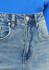 Light blue denim jeans with faded areas, five-pocket design, metal button closure, and contrast stitching. Textured cotton fabric.