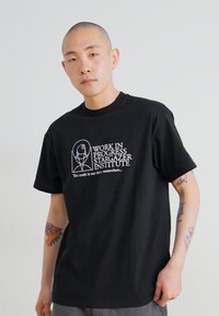 T-shirt preto de manga curta com texto e gráfico brancos: "OBRA EM ANDAMENTO INSTITUTO DE OBERVAÇÃO DAS ESTRELAS" com um design circular e um slogan abaixo.