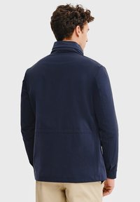 Granatowa kurtka z wysokim kołnierzem, o gładkim, matowym wykończeniu. Projekt obejmuje długie rękawy i szew na plecach.