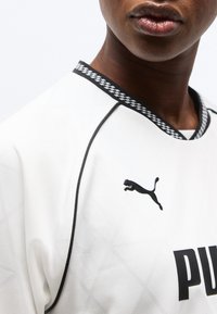 Κοντινό πλάνο ατόμου που φοράει λευκό αθλητικό μπλουζάκι Puma με μαύρα περιθώρια και λογότυπο στο στήθος, δείχνοντας το κάτω μέρος του προσώπου και τον λαιμό.