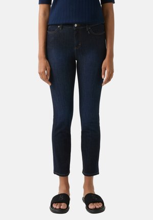 Dark blue denim jeans with a slim fit, featuring a zip fly, belt loops, and contrasting stitching. Paired with black slide sandals.