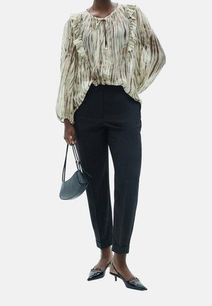 Femme portant un chemisier beige à volants et motifs, un pantalon noir tailleur, des chaussures noires à talons bas, tenant un petit sac à main noir.