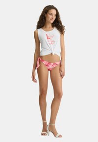 Top bianco senza maniche con orlo annodato e grafica rosa. Slip bikini con stampa tropicale rosa e lacci laterali. Indossato con sandali bianchi con tacco.