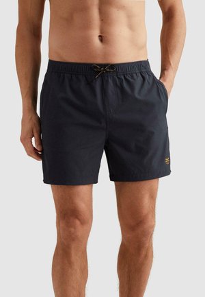 Close-up of a man wearing black drawstring swim shorts with pockets, standing against a plain light background.