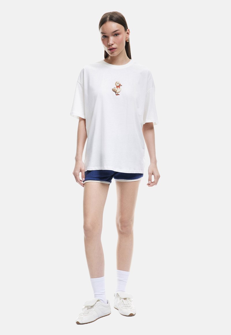 Camiseta blanca de algodón con mangas cortas y escote redondo, que presenta un pequeño diseño de personaje bordado en el pecho. Combinada con pantalones cortos azules.