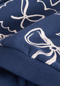 Tissu en coton marine avec imprimés de fleurs blanches et de rubans ; ourlet côtelé ajoutant de la texture. Vue en gros plan mettant en valeur les détails du design.