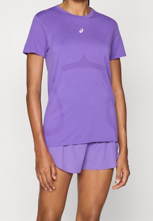 Osoba w sportowej, fioletowej koszulce z krótkim rękawem z logo i pasujących szortach, stojąca na tle jednolitego, jasnego tła.