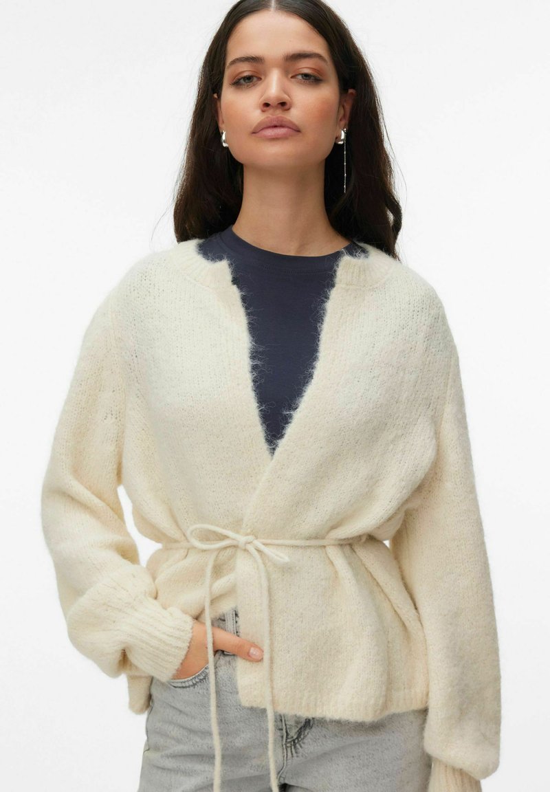 Cardigan en maille couleur crème à manches longues, devant ouvert et ceinture à nouer. Présente une texture douce et légèrement duveteuse ainsi qu'une coupe décontractée.