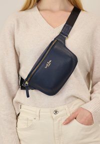 Marsupio in pelle blu navy con zip dorata, manico superiore e tracolla regolabile, indossato a tracolla sopra un leggero maglione e pantaloni crema.