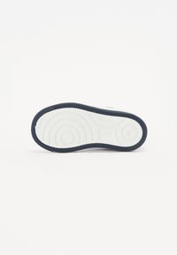 Semelle de sneaker blanche avec un bord en caoutchouc de couleur marine texturé, présentant des motifs circulaires et des rainures pour l'adhérence. Surface lisse sur la partie supérieure.
