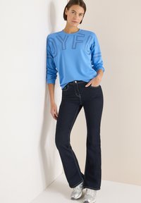 Jasnoniebieska bluza z napisem „JOYFUL” w ciemnych konturach, zestawiona z ciemnoniebieskimi dzwonami z kontrastującymi przeszyciami oraz srebrnymi tenisówkami.