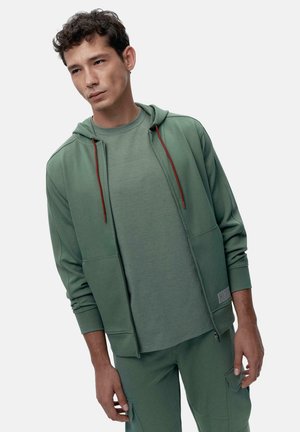 Felpa verde con lacci rossi, abbinata a una maglietta verde coordinata, presenta una vestibilità comoda e una superficie liscia. Abbigliamento atletico informale.