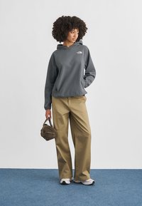 Γκρι φούτερ με κουκούλα και ριμπ υφή, χαλαρή εφαρμογή; μπεζ παντελόνι με φαρδύ πόδι; κρατώντας μια μικρή καφέ τσάντα; λευκά αθλητικά παπούτσια.