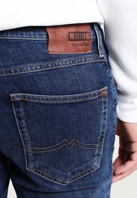 Mörkblå denimjeans med en brun läderlapp med texten "MUSTANG True Denim." Designen har två bakfickor med kontrasterande sömmar.