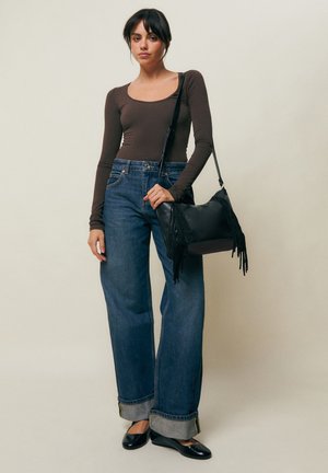 Žena v priliehavom hnedom top s dlhým rukávom, modrých širokých nohaviciach s manžetami, čiernych balerínach a nesie čiernu kabelku s strapcami na rameno.