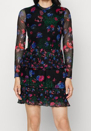 Femme portant une robe noire transparente à manches longues avec des motifs floraux et d’oiseaux colorés et une jupe à volants superposés.