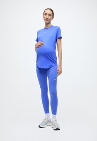 Μπλε αθλητικό τοπ εγκυμοσύνης και κολάν από ελαστικό ύφασμα· διαθέτει λογότυπο στο στήθος, σε συνδυασμό με ανοιχτό γκρι αθλητικά παπούτσια.