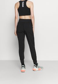 Schwarze Sporthose und passendes ärmelloses Oberteil. Die Hose hat einen elastischen Bund und verjüngte Bündchen. Die Schuhe sind grau mit orangefarbenen Akzenten.