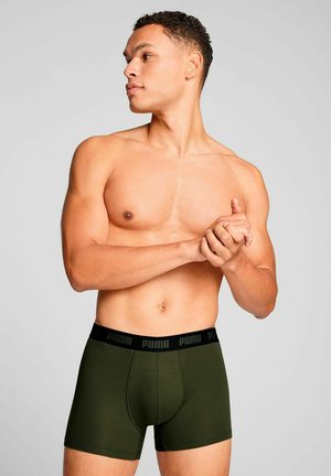 Dunkelgrüne Boxer-Shorts aus dehnbarem Material, mit einem schwarzen Bund, der das Logo "PUMA" trägt. Das Modell posiert mit gefalteten Händen.