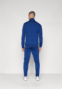 Modra trenirka s ovratnikom, belimi stranicami in rebrastimi manšetami. Slim-fit oblikovanje, izdelana iz gladkega blaga, prikazana s hrbtne strani.