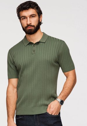 Mężczyzna z brodą w zielonym prążkowanym polo z krótkim rękawem i zegarkiem na rękę, z jedną ręką w kieszeni ciemnych jeansów.