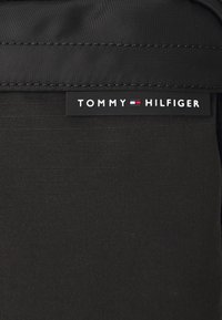 Fekete szintetikus táska, amelyen nagy fehér "TOMMY HILFIGER" logó címke található vörös és kék részletekkel. Simább textúra enyhe fényességgel.