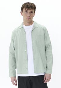 Light green button-up shirt made of textured fabric with a collar, single chest pocket, and white t-shirt underneath. Black pants worn.