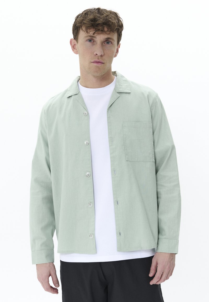 Light green button-up shirt made of textured fabric with a collar, single chest pocket, and white t-shirt underneath. Black pants worn.