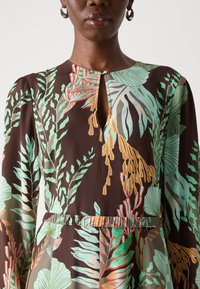 Abito a fantasia floreale marrone, con stampe di foglie verdi, rosse e beige. Scollo a chiave, maniche lunghe e dettaglio vita arricciata.